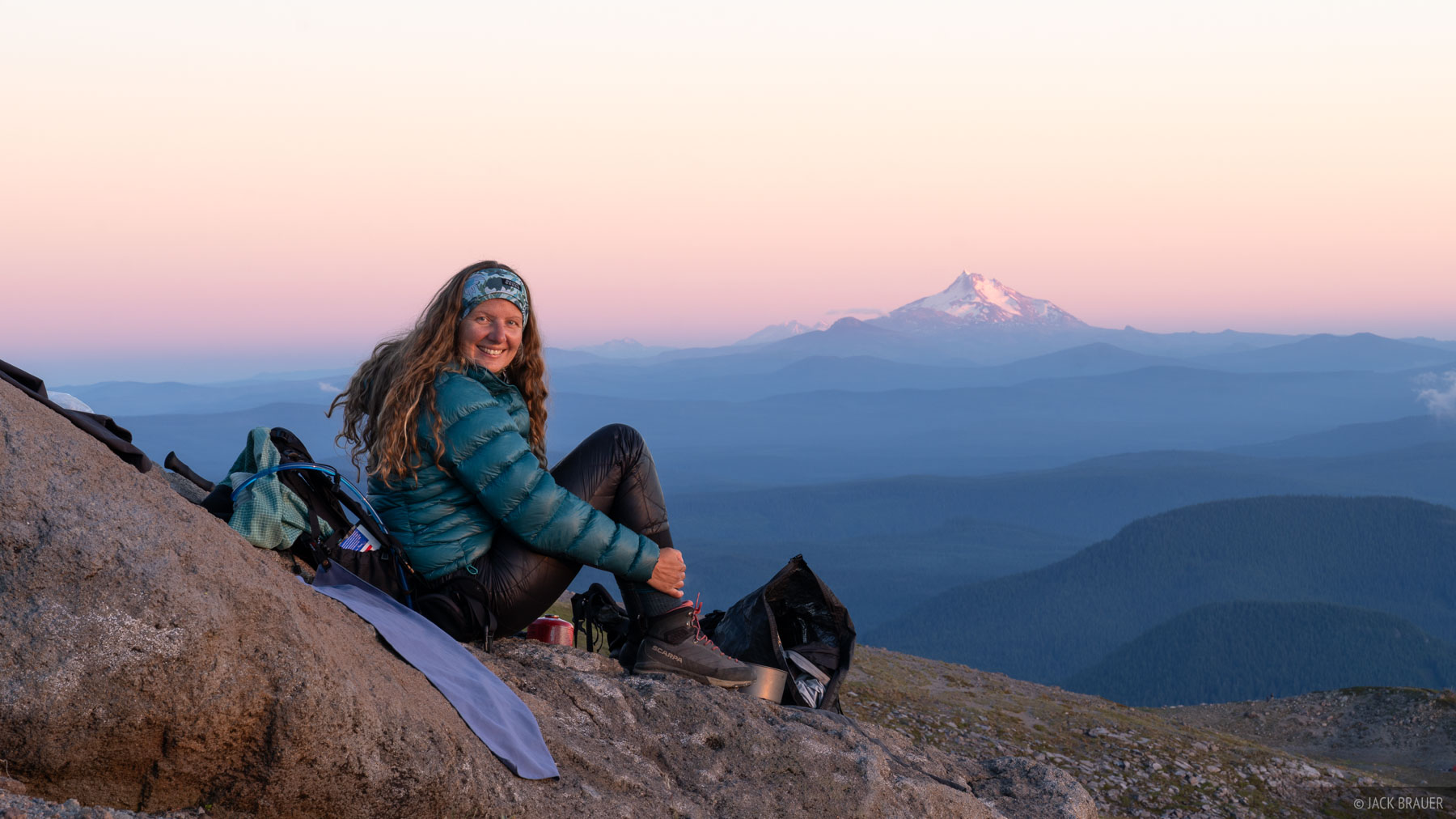 A hiker sits on Mount Hood with Mount Jefferson in the background at sunset.